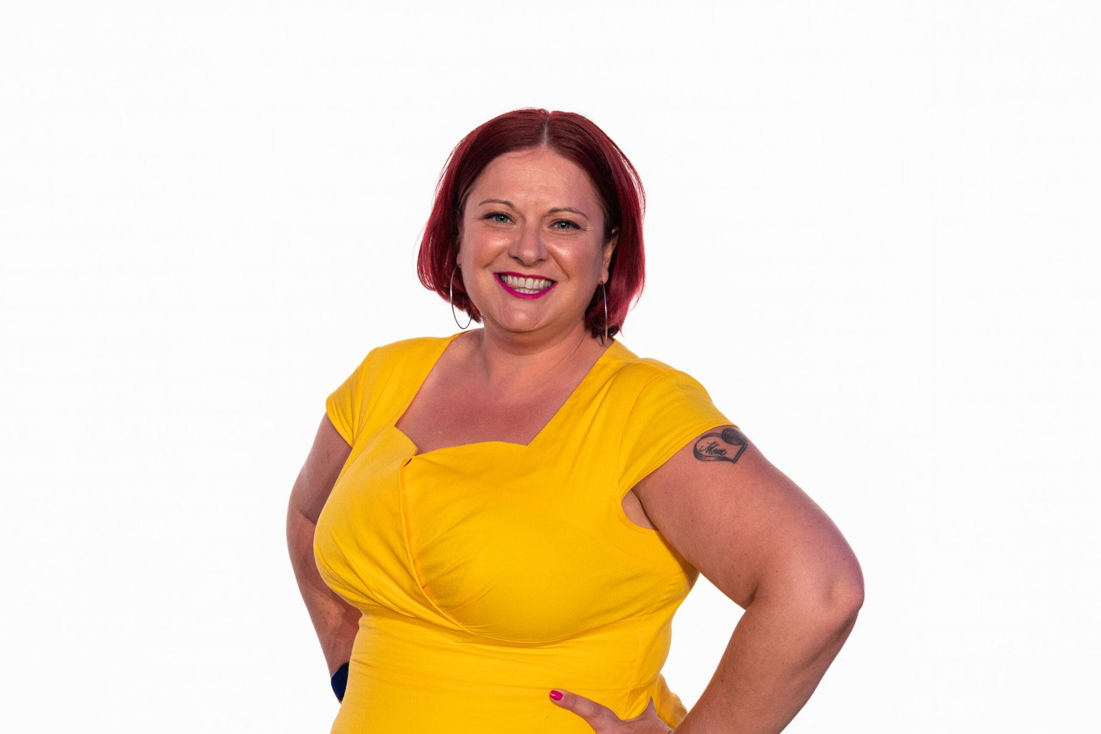 Britne Jenke, a white woman with red hair, smiles in pink lipstick with her hands on her hips. She is wearing a yellow dress and a heart tattoo is visible on her left arm.