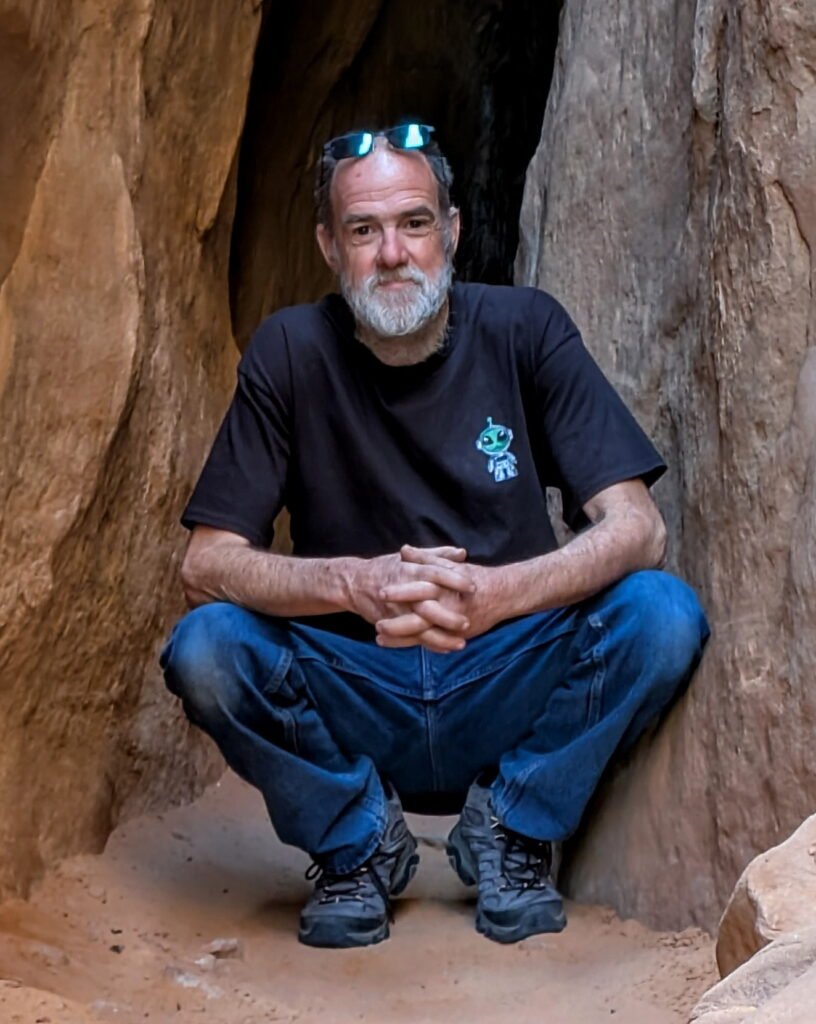 Michael Bodman, a white slender man with grey hear and beard, wearing a black tshirt and jeans, squatting with his hands folded in front of a small rocky cave.