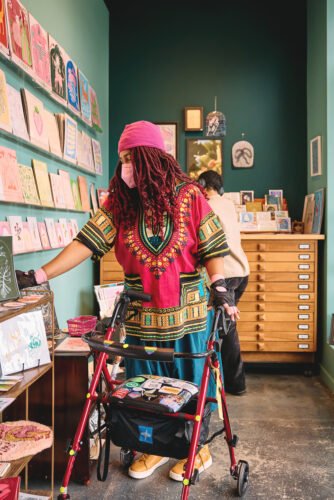 Two friends look through art prints in a gift shop. A masked Black non-binary woman reaches towards a print, with one hand remaining on their rollator. They’re in bright colors, including a beanie hat, dashiki, harem pants, and sneakers. Their hands are layered with bike gloves over arthritis gloves and their rollator is covered in stickers. In the background, a Black non-binary person can be seen from the back, wearing noise-canceling headphones.
