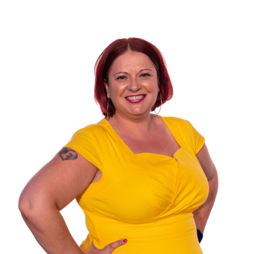 Britne Jenke, a white woman with red hair, smiles in pink lipstick with her hands on her hips. She is wearing a yellow dress and a heart tattoo is visible on her left arm.